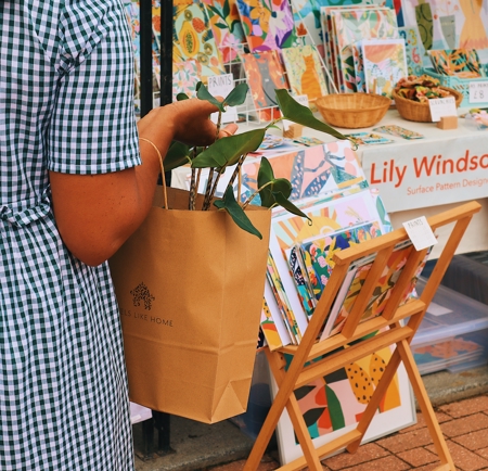 Person wearing a green-and-white checkered dress holding a brown paper bag with a leafy plant inside, standing in front of a market stall displaying colorful patterned prints and artwork. A wooden stand holds more prints, and a sign on the table reads ‘Lily Windsor Surface Pattern Design.’