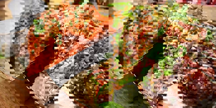 Close-up of two trays of saucy chicken wings served on a wooden table. The tray in the foreground features wings coated in a sticky glaze, topped with sesame seeds, chopped green onions, and a drizzle of creamy sauce, with a lime wedge on the side. The tray in the background contains wings covered in bright orange sauce, garnished with finely chopped herbs. Two small white dipping cups are placed alongside the wings.