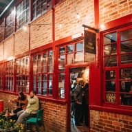 The red brick exterior of Hop Hideout at Leah's Yard, with red painted windows and doors and a black hanging sign that says 'Hop Hideout'. A man and a woman are sat on a table outside drinking, the door is open and customers are waiting at the bar.