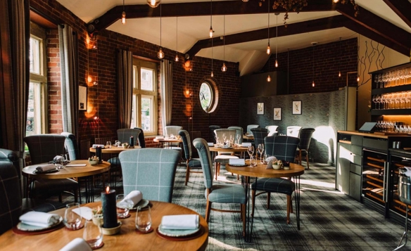An elegant restaurant interior featuring round wooden tables set with folded napkins, glassware, and small decorative items. The space has exposed brick walls, large windows with gray curtains, and a patterned carpet. Pendant lights hang from the ceiling beams, creating warm ambient lighting. A wine storage area and shelving with glasses are visible on the right side of the room, adding to the sophisticated atmosphere.