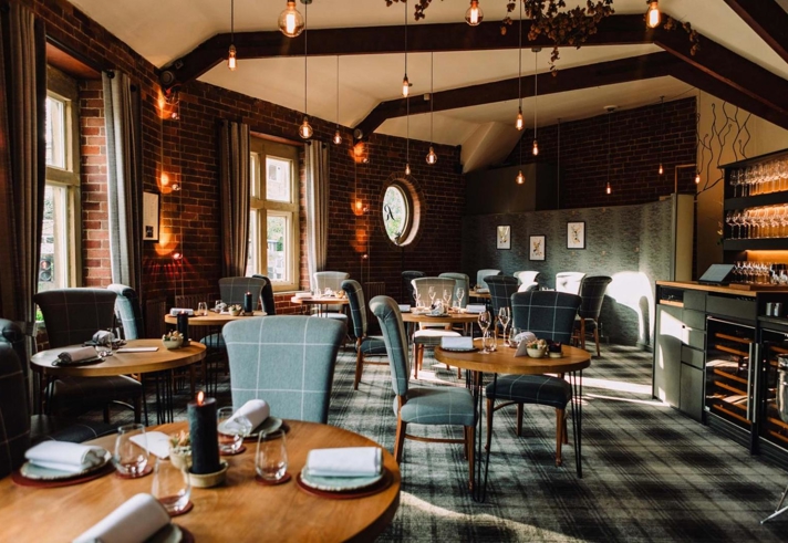 An elegant restaurant interior featuring round wooden tables set with folded napkins, glassware, and small decorative items. The space has exposed brick walls, large windows with gray curtains, and a patterned carpet. Pendant lights hang from the ceiling beams, creating warm ambient lighting. A wine storage area and shelving with glasses are visible on the right side of the room, adding to the sophisticated atmosphere.