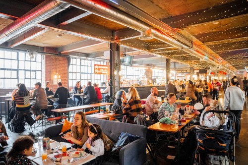 A lively indoor food hall with long wooden tables and benches filled with people eating and drinking. The space has exposed brick walls, large industrial-style windows, and string lights hanging from the ceiling, creating a warm and festive atmosphere. A stroller is visible in the foreground, and the hall extends into the background with more diners.