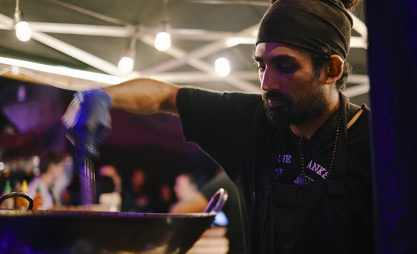 A person wearing a dark apron and head wrap is cooking at an outdoor food stall under a canopy with string lights. The individual is stirring food in a large wok, and bottles of condiments are visible in the background along with blurred figures and warm lighting.