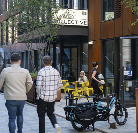 Two people walking along a paved pedestrian street lined with modern buildings featuring large windows and rust-colored panels. In the foreground, a cargo-style electric bike is parked near a planter. To the right, a café with bright yellow chairs and tables is visible, with one person seated and another carrying drinks. A sign above the café entrance reads “COLLECTIVE.” Trees and steps leading to an upper level appear in the background.