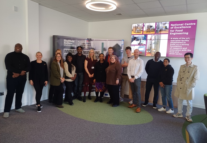 A group of twelve people standing in a semi-circle inside a modern meeting room with grey carpet and green accents. Behind them is a large banner displaying “Sheffield Hallam University” and “National Centre of Excellence for Food Engineering.” On the right wall, there is a digital screen showing images and text about the centre. The room has white walls, a ceiling light, and a few chairs visible on the side.