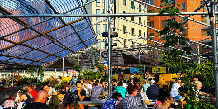  A busy outdoor seating area filled with people at wooden benches under a transparent canopy. The sun shines brightly overhead, casting strong light and highlighting surrounding greenery.