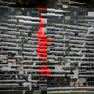 Panoramic view of a packed football stadium with fans filling the stands. The green pitch is set for a match, with goalposts visible at both ends. Large red banners and flags are displayed by supporters, and the stadium roof arches overhead with a modern design.