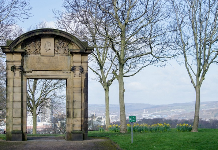 A historic stone archway with decorative carvings stands in a park surrounded by bare trees. The arch frames a distant view of a cityscape with hills in the background. Yellow daffodils bloom in the grassy area near the arch, and a small green sign is visible on the lawn. The sky is bright with scattered clouds, creating a clear and open atmosphere.