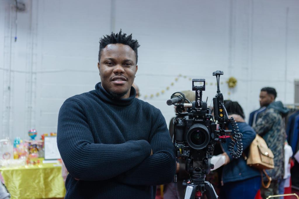 A person wearing a dark sweater stands with arms crossed next to a professional video camera mounted on a tripod. The camera has various attachments, including a microphone and monitor. In the background, there are people, colourful decorations, and tables covered with bright cloths, suggesting an indoor event or gathering.