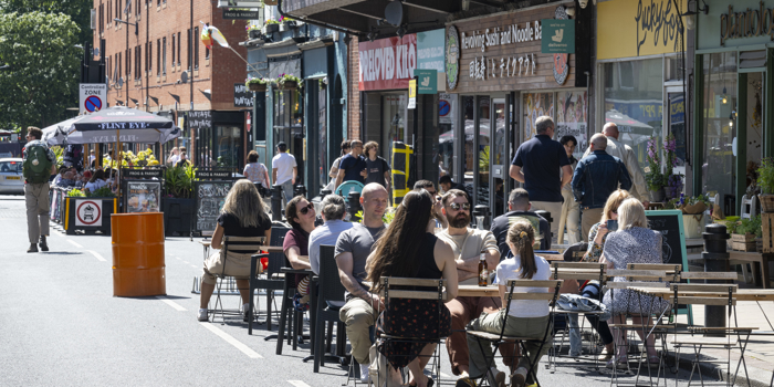 People are seated at outdoor tables on a closed-off city street, enjoying food and drinks in sunny weather. The street is lined with shops and restaurants, including signs for a noodle bar and a café. A bright orange barrel and potted plants decorate the area, while pedestrians walk along the street in the background. The scene conveys a lively, social atmosphere in an urban setting.