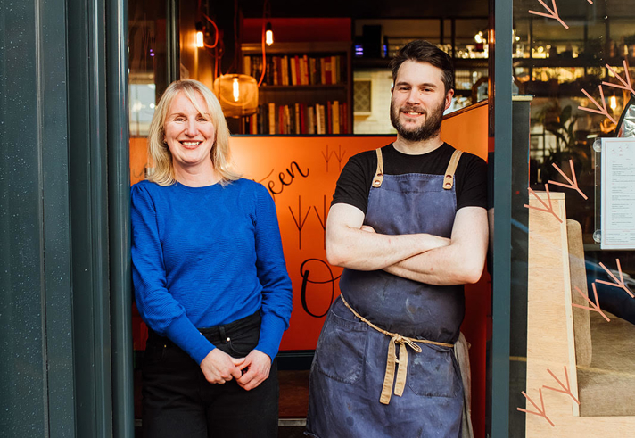 Two people standing in the doorway of a cozy restaurant or café. One is wearing a bright blue long-sleeve top and dark pants, while the other is dressed in a black shirt with a dark apron tied at the waist. The interior behind them features warm lighting, shelves with books, and string lights. A handwritten orange sign is partially visible inside, and the exterior has dark framing around the entrance.