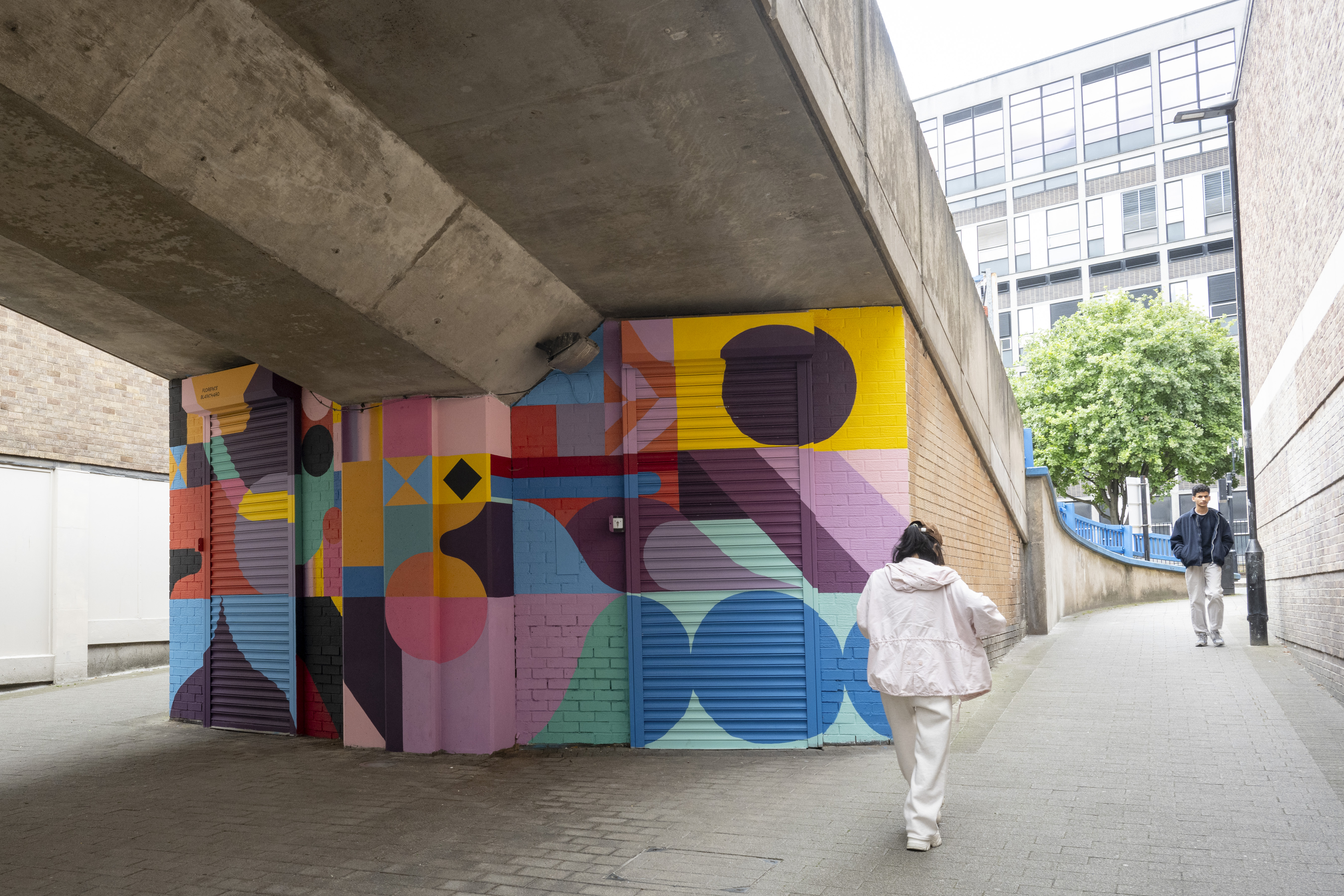 Urban walkway beneath a concrete overpass featuring a vibrant geometric mural with bold shapes and bright colors including yellow, pink, blue, and purple. Two people are walking along the path, and modern buildings and greenery are visible in the background