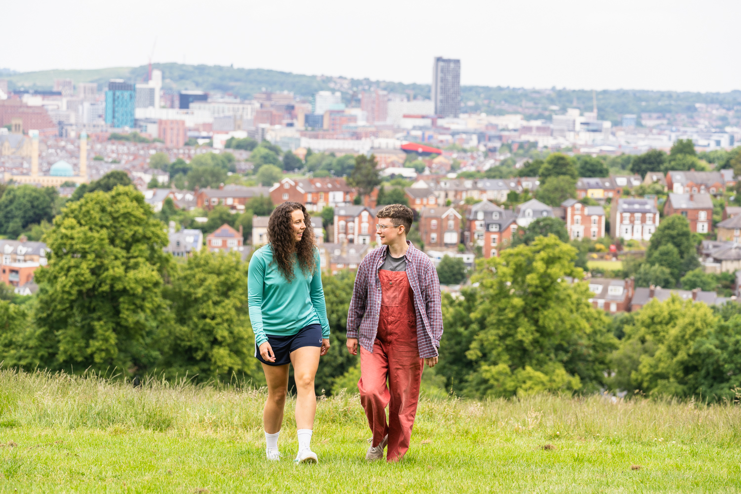 A couple walking through Meersbrook Park with green lawns, tress and a view of the Sheffield cityscape in the background 