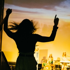 A silhouette of a person with arms raised, standing in front of a brightly lit stage where musicians are performing. The background features a large screen displaying a vivid sunset scene with orange and yellow hues. On stage, a drum set and keyboard are visible, with performers seated behind the instruments. The image captures the lively atmosphere of a concert or music event indoors.