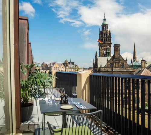 Outdoor balcony with a small dining table set for two, overlooking a historic cityscape with a tall clock tower and other ornate buildings under a partly cloudy blue sky.