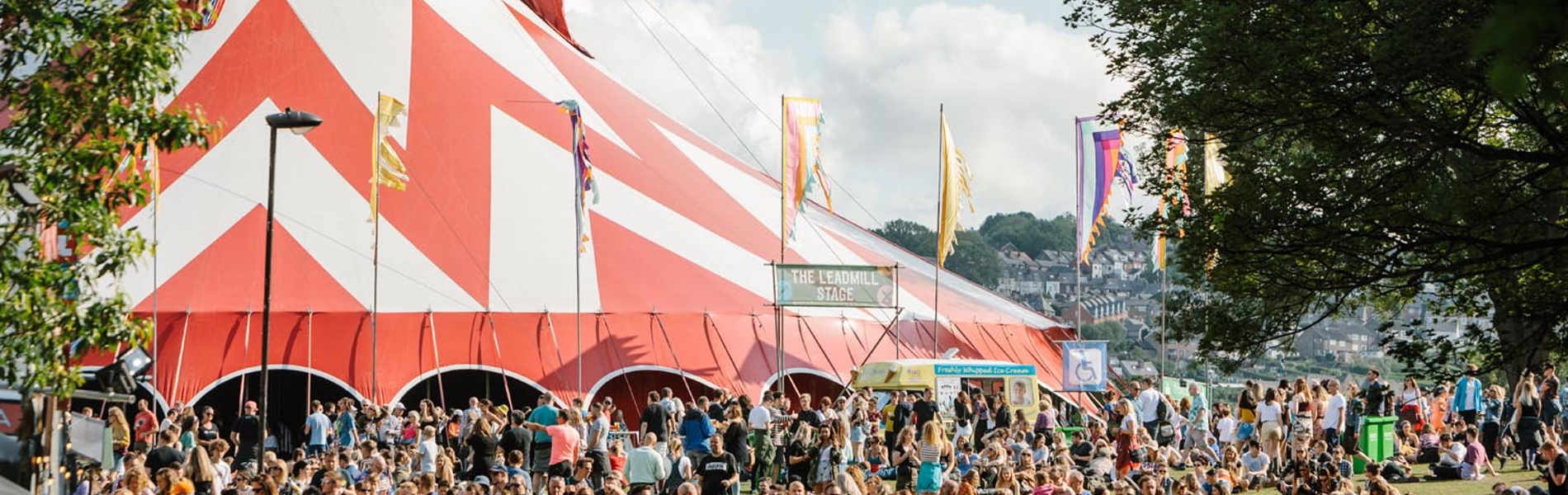 Large outdoor festival crowd gathered near a big red-and-white striped tent, with colourful flags and trees in the background under a partly cloudy sky.