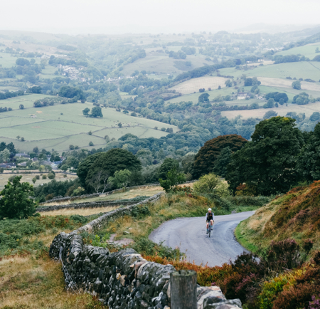 Aerial shot of cyclist riding along countryside road with a stone wall, further fields of countryside in the background.