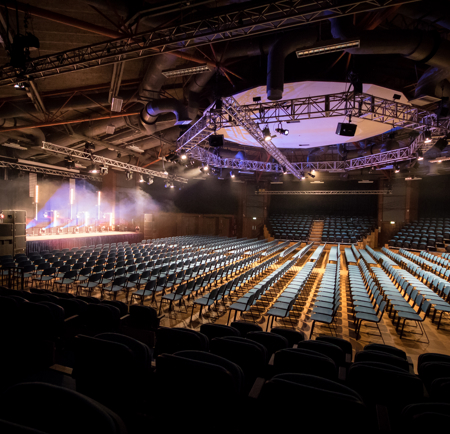 Large auditorium set up in theatre style with rows of blue chairs facing a stage. The stage is illuminated with bright spotlights and features podiums and equipment. Overhead, a circular truss with lighting rigs hangs from the exposed industrial ceiling with beams and ducts. The space is dimly lit except for the stage area, creating a dramatic atmosphere.