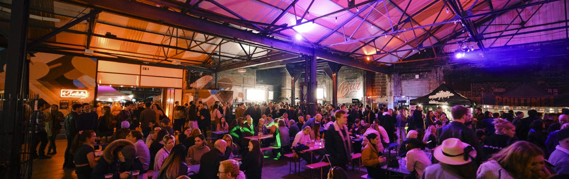 Large indoor venue with high industrial-style ceiling and exposed beams, filled with people seated at long wooden tables and standing in groups. Warm orange and purple lighting creates a lively atmosphere, with food and drinks visible on tables. A bar and food stalls are in the background.