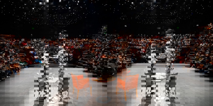 Two speakers standing on a stage in front of a full audience inside a theatre with star‑like ceiling lights.