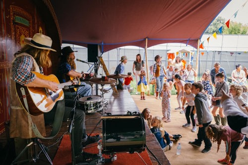 A lively scene under a large tent where two musicians are performing on a small stage. One musician is playing an acoustic guitar while the other is seated at a drum set. In front of the stage, a group of children and adults are gathered, some dancing and others watching the performance. The area is decorated with colorful bunting, and the background shows outdoor structures and greenery, suggesting a festival or community event.