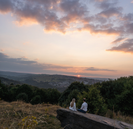 Two people sitting on a large flat rock in a grassy hillside area, overlooking a valley with scattered trees and distant houses. The sky is filled with soft clouds illuminated by the warm colours of a sunset, casting a serene glow across the landscape. Yellow wildflowers are visible in the foreground.