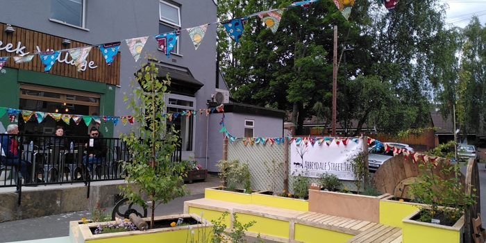 Outdoor seating area with bright yellow and beige wooden benches and planters containing small trees and plants. Colorful triangular bunting is strung overhead, and a banner reading ‘Abbeydale Street’ is visible on a fence. A café with a green storefront and people sitting outside is in the background.