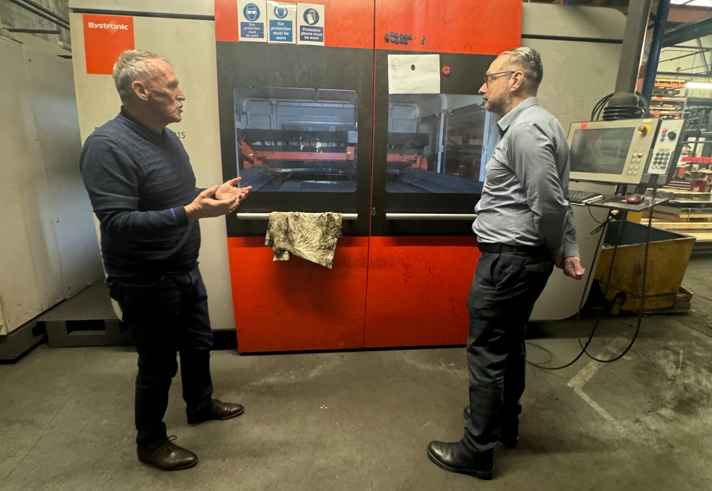 Two people standing in front of a large industrial machine with a bright orange and black exterior in a factory setting. One person is gesturing with hands while the other stands near a control panel attached to the machine. The background shows metal beams, shelves, and equipment under a high roof with skylights.