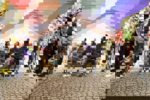 Three people sitting on a yellow sofa under a large Progress Pride flag being held overhead by a group of people. The flag displays rainbow colors along with chevron stripes in black, brown, white, pink, blue, and light blue. The scene takes place outdoors on a cobblestone walkway, with individuals standing around holding sections of the flag.