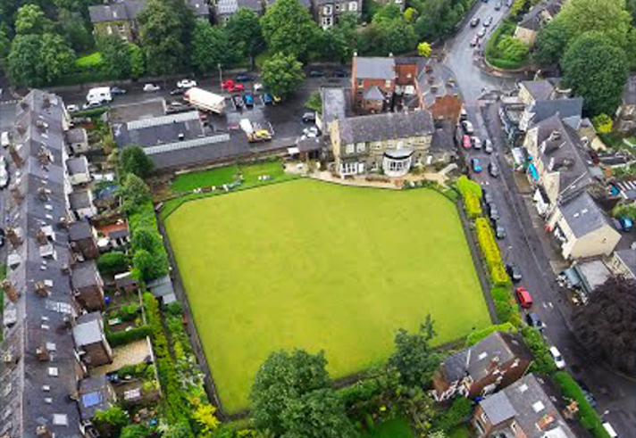 Aerial view of a rectangular green lawn, possibly a bowling green, surrounded by residential houses and narrow streets. The area includes trees, gardens, and parked cars, with a mix of brick buildings and greenery visible throughout the neighbourhood.