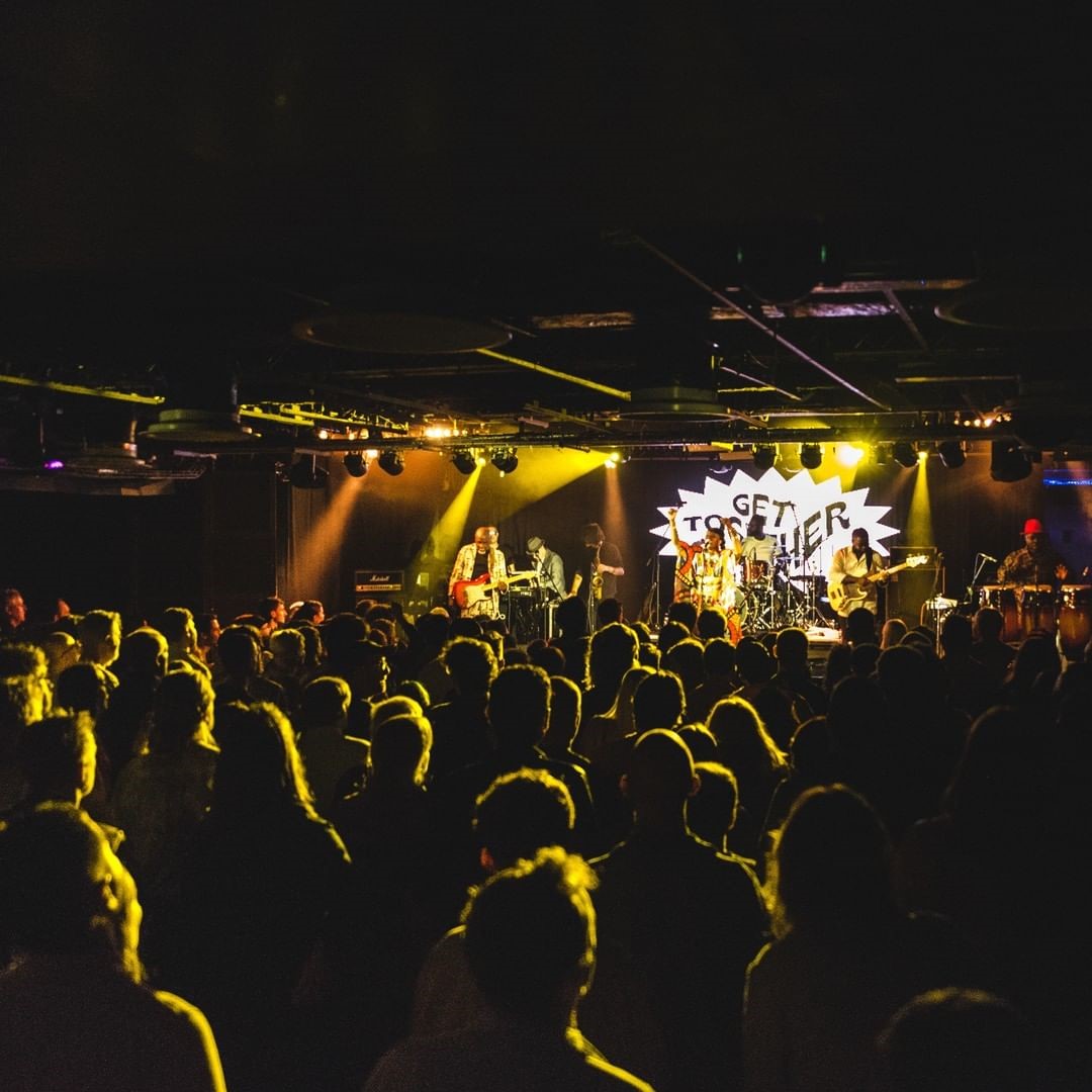 A large crowd of people is gathered in a dimly lit venue, watching a live band perform on stage. The stage is illuminated with yellow lights, and the backdrop features a sign that reads "GET TOGETHER" in bold letters. The band consists of multiple musicians playing various instruments, including guitars and drums. The audience appears engaged with the performance.