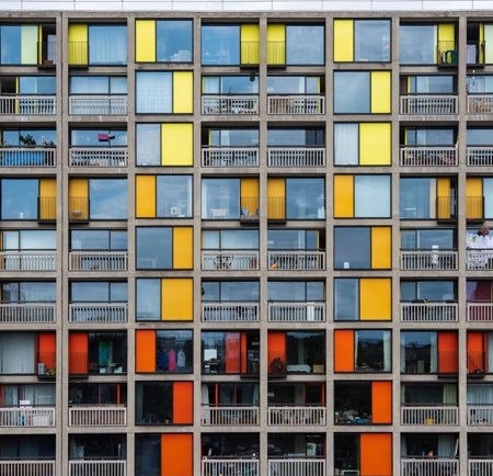 Front view of a multi-story residential building with a grid-like facade featuring large windows and panels in shades of yellow, orange, and red. Each unit has a balcony with railings, and some balconies display hanging laundry or personal items.