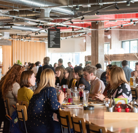 Kommune Food Hall - Interior of a modern food hall with industrial-style design, featuring exposed ducts, concrete pillars, and large windows. Several groups of people are seated at long wooden tables enjoying food and drinks. The space is well-lit with natural light and hanging ceiling lights, and a black menu board is visible in the background.