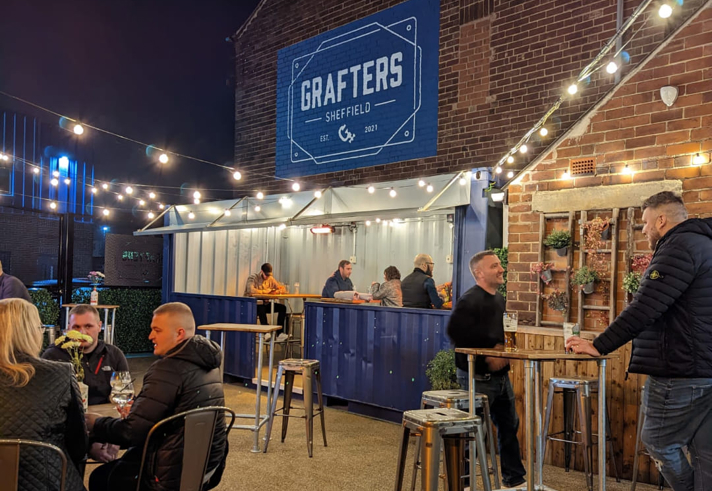 An outdoor bar area at night with string lights overhead, people sitting and standing at high tables, and a blue “Grafters Sheffield” sign mounted on a brick wall above a serving counter.