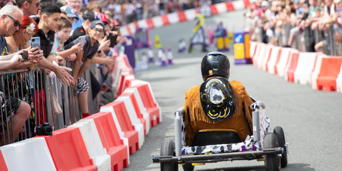 A soapbox cart with a simple frame and four wheels races down a city street lined with red-and-white barriers. The driver wears a black helmet and a fringed brown jacket with a decorative design on the back. Spectators crowd behind metal railings on both sides, watching the event. Purple-and-white race markers and colourful props are visible further down the track.