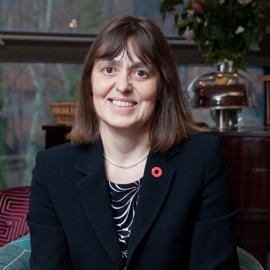 A person dressed in a black blazer with a patterned top underneath, seated indoors. The blazer has a red poppy pin on the lapel, and the individual is wearing a pearl necklace. The background features a glass window, a plant arrangement, and decorative items on a wooden surface.