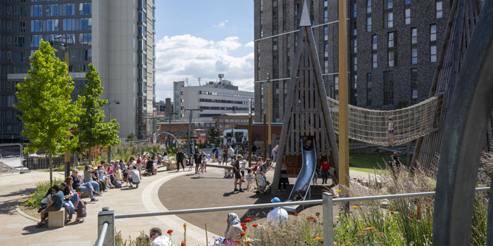 An urban playground surrounded by tall modern buildings, with people sitting on curved benches and children playing on wooden climbing structures and slides. The area includes a rope bridge, landscaped flower beds with colorful blooms, and paved walkways. The background shows city architecture under a bright blue sky with scattered clouds.
