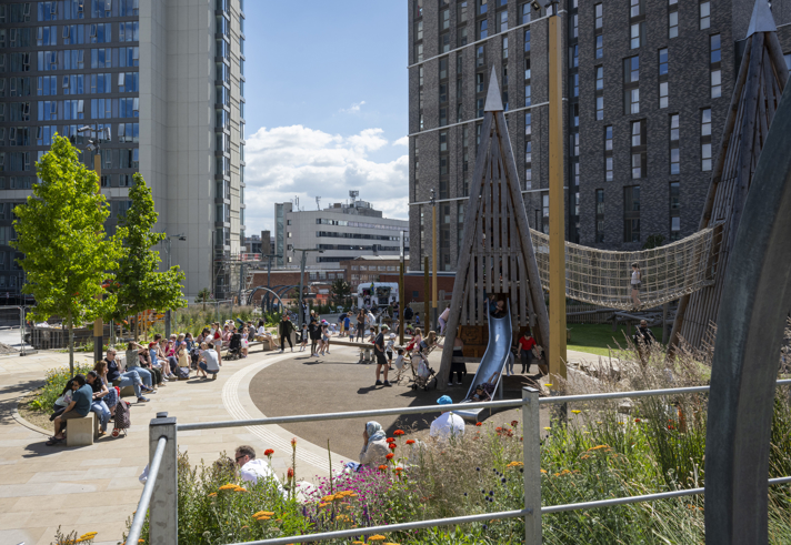 An urban playground surrounded by tall modern buildings, with people sitting on curved benches and children playing on wooden climbing structures and slides. The area includes a rope bridge, landscaped flower beds with colorful blooms, and paved walkways. The background shows city architecture under a bright blue sky with scattered clouds.