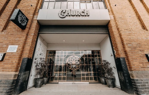 Entrance to a building with a sign reading “Church – Temple of Fun” above the doorway. The entrance features decorative gold metalwork on glass doors, flanked by potted plants. The building has a brick exterior, and a black hanging sign with a white graphic is visible on the left side.