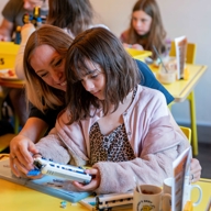 Two people sit at a bright yellow table in a café, assembling a toy train model. Other tables with children and adults are visible in the background, along with mugs and menus on the table, creating a playful and family-friendly atmosphere.