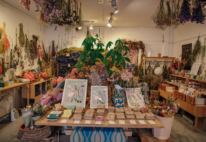 Interior of a boutique shop filled with dried flowers, plants, and botanical arrangements. A central wooden table displays illustrated floral prints, seed packets, and small items, while shelves and walls around the room are decorated with hanging bunches of dried flowers and greenery. The space has warm lighting and a natural, rustic aesthetic.