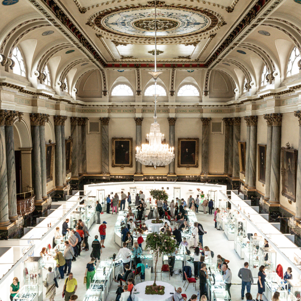 Elegant exhibition inside a grand hall with ornate columns, arched windows, and a decorative ceiling featuring a large chandelier. Visitors browse display booths arranged in a square layout around a central tree.