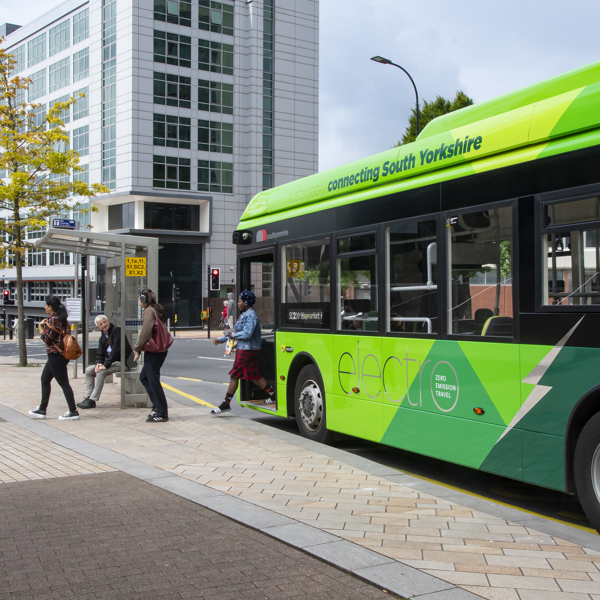 A bright green electric bus with the text “connecting South Yorkshire” on the side is stopped at a bus stop in an urban area. Several people are boarding the bus from a paved sidewalk. The background features modern multi-story office buildings with large glass windows, street signs, and a tree with yellow leaves under a partly cloudy sky.